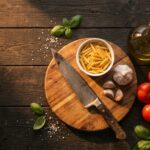 Essentiels cuisine débutant : couteau de chef, pâtes, ail frais, tomates, oeufs et huile d'olive sur planche en bois